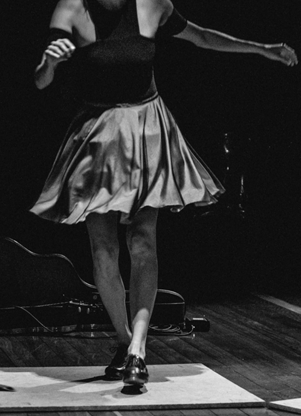 Black and white photo of a tap dancer standing on a small square wooden platform. She wears black tights, silver skirt and black shoes. Behind the tap dancer, a ukulele case lying on the stage.