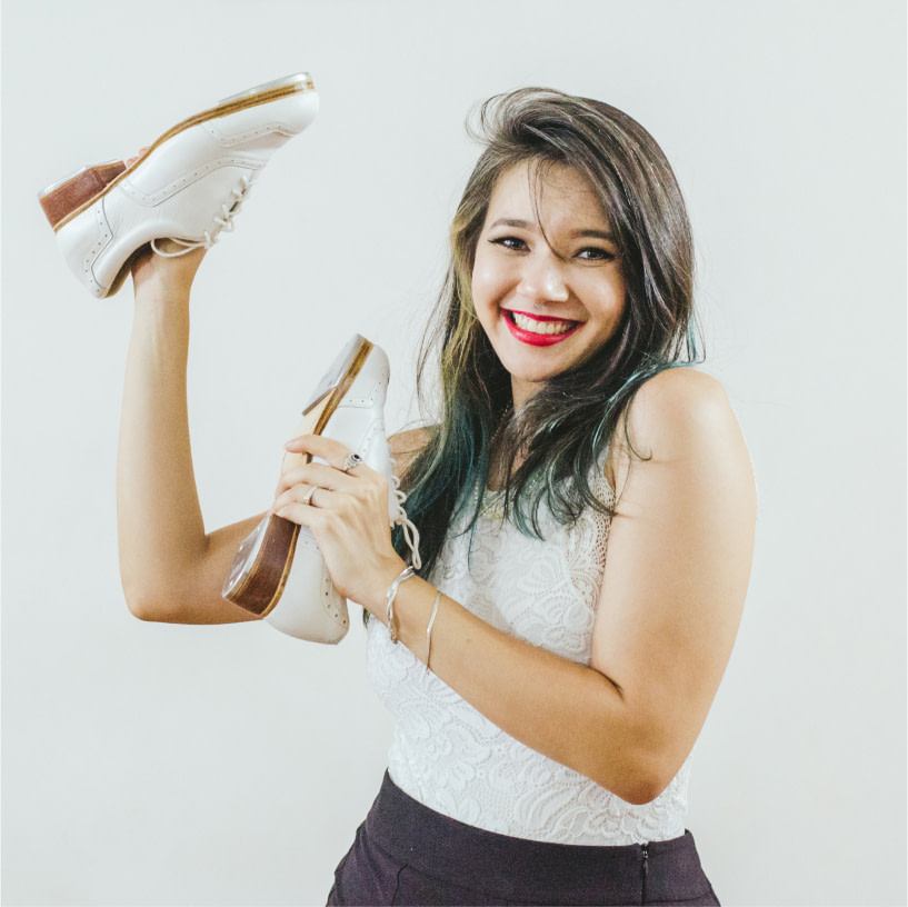 Foto colorida de Ana Luiza, sapateadora do elenco, sorridente, segurando um par de sapatos de sapateado brancos, ao lado do rosto.