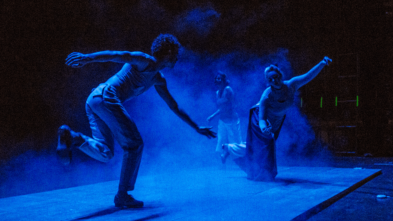 Color photo of three tap dancers dancing on stage, with blue stage lighting and smoke. Rodrigo is on his side with his left leg bent and off the ground as he leans his body forward with his arms outstretched. Ana is in the front doing the same move as Rodrigo while Renata is standing further back preparing for her next move.