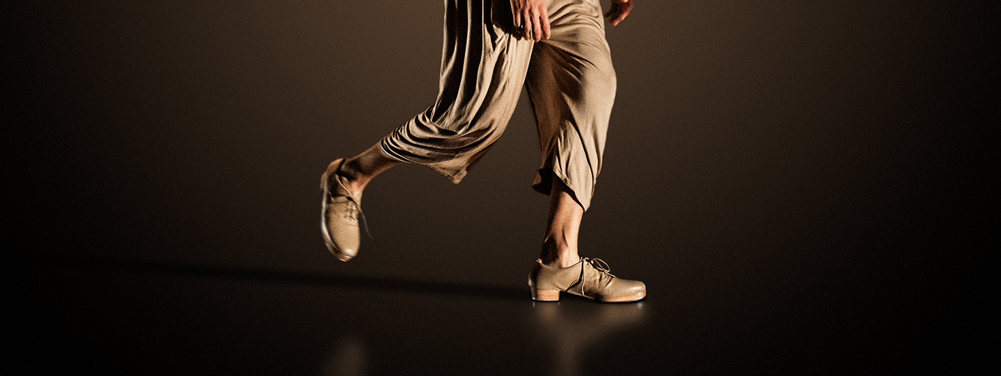 Imagem de Rodrigo, sapateador do elenco, vestindo macacão na cor bege e sapatos de sapateado na mesma cor. O foco da imagem está em suas pernas e pés: um pé apoiado no chão e o outro no ar, apontando para trás.