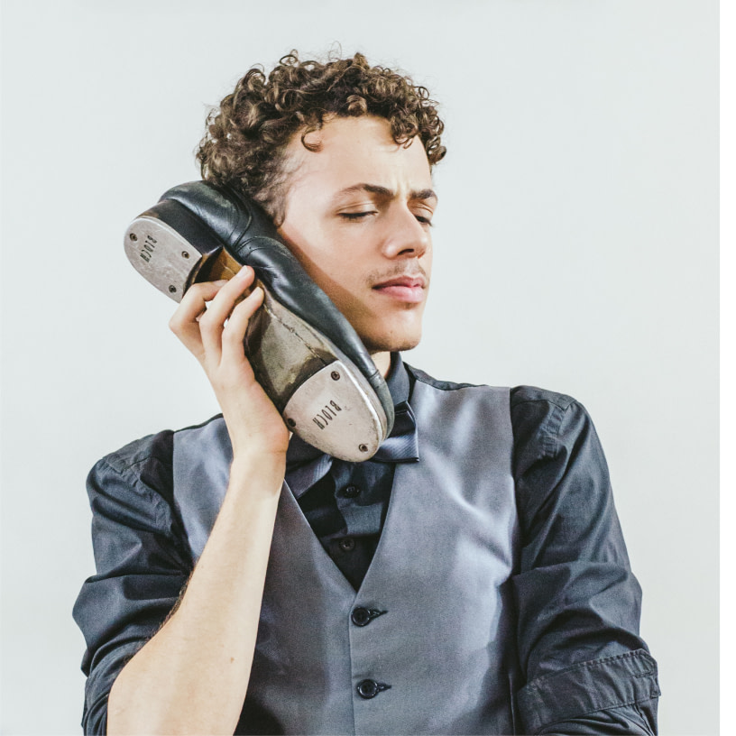Color photo of Rodrigo, the cast tap dancer, with a looking thoughtful, holding a tap shoe close to his right ear.