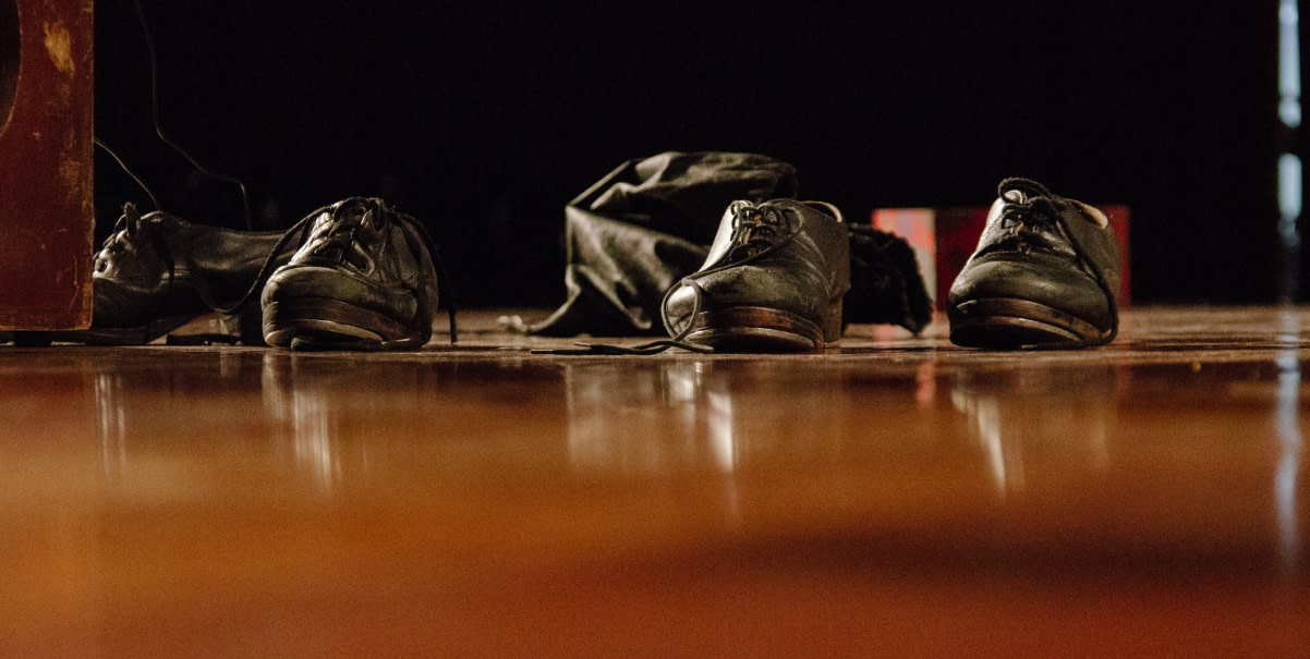 Foto colorida de dois pares de sapato de sapateado na cor preta, dispostos em cima do palco de madeira. Na lateral esquerda, é possível notar uma parte do instrumento percussivo “cajón”.